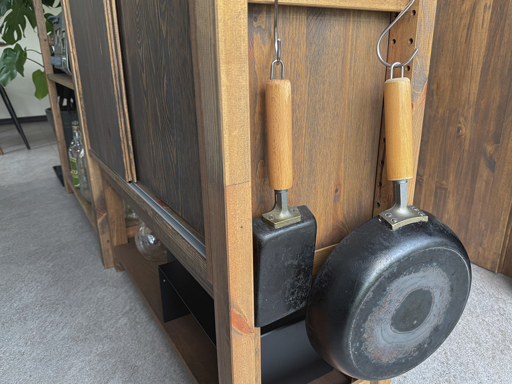 木製棚の側面にS字フックを使い、鉄製のフライパンと玉子焼き器を吊るして収納したクローズアップ